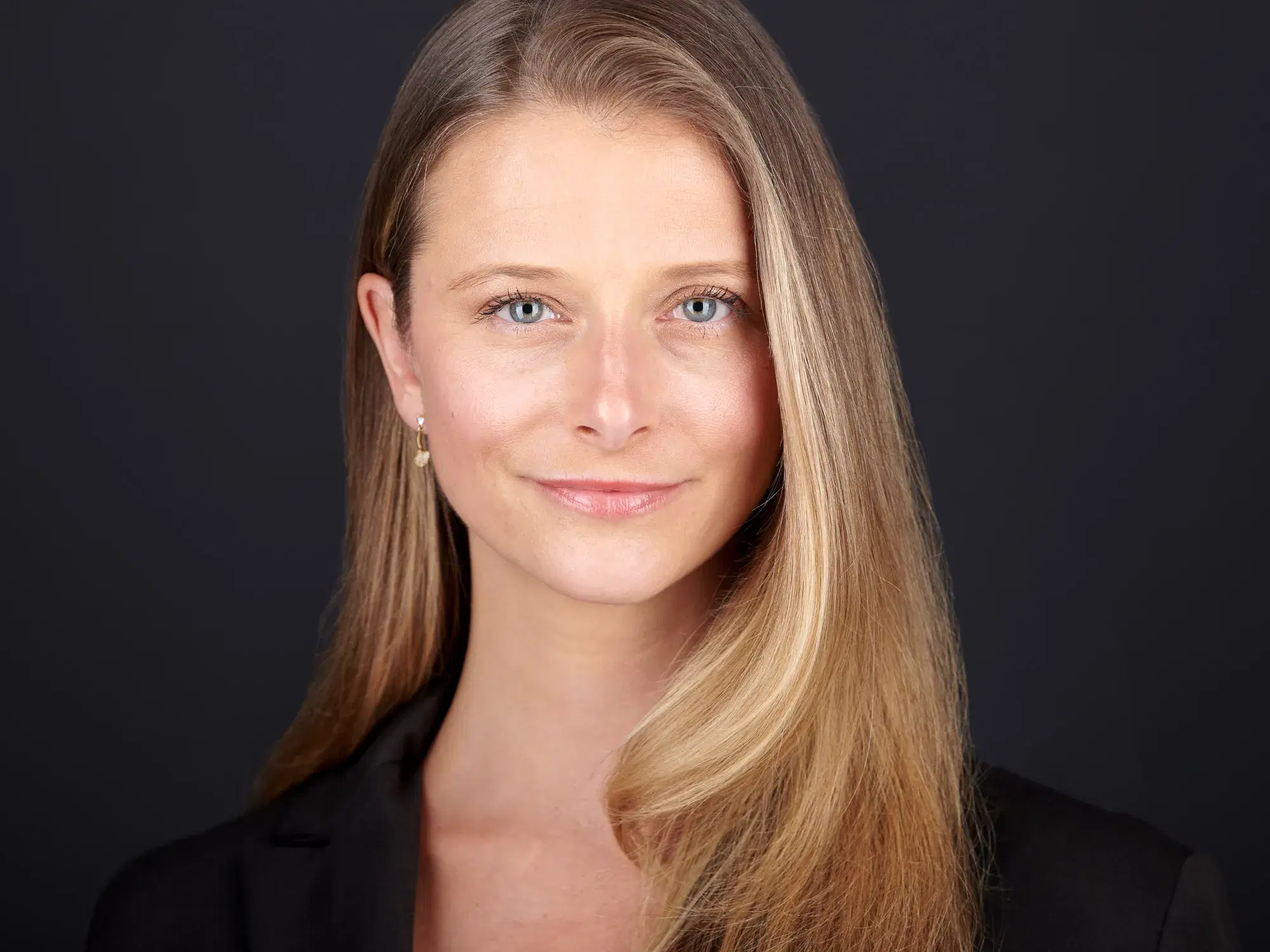Professional headshot of young woman with long blonde hair and blue eyes in black top against dark studio backdrop — Boston team headshots by Damon Bates Photography
