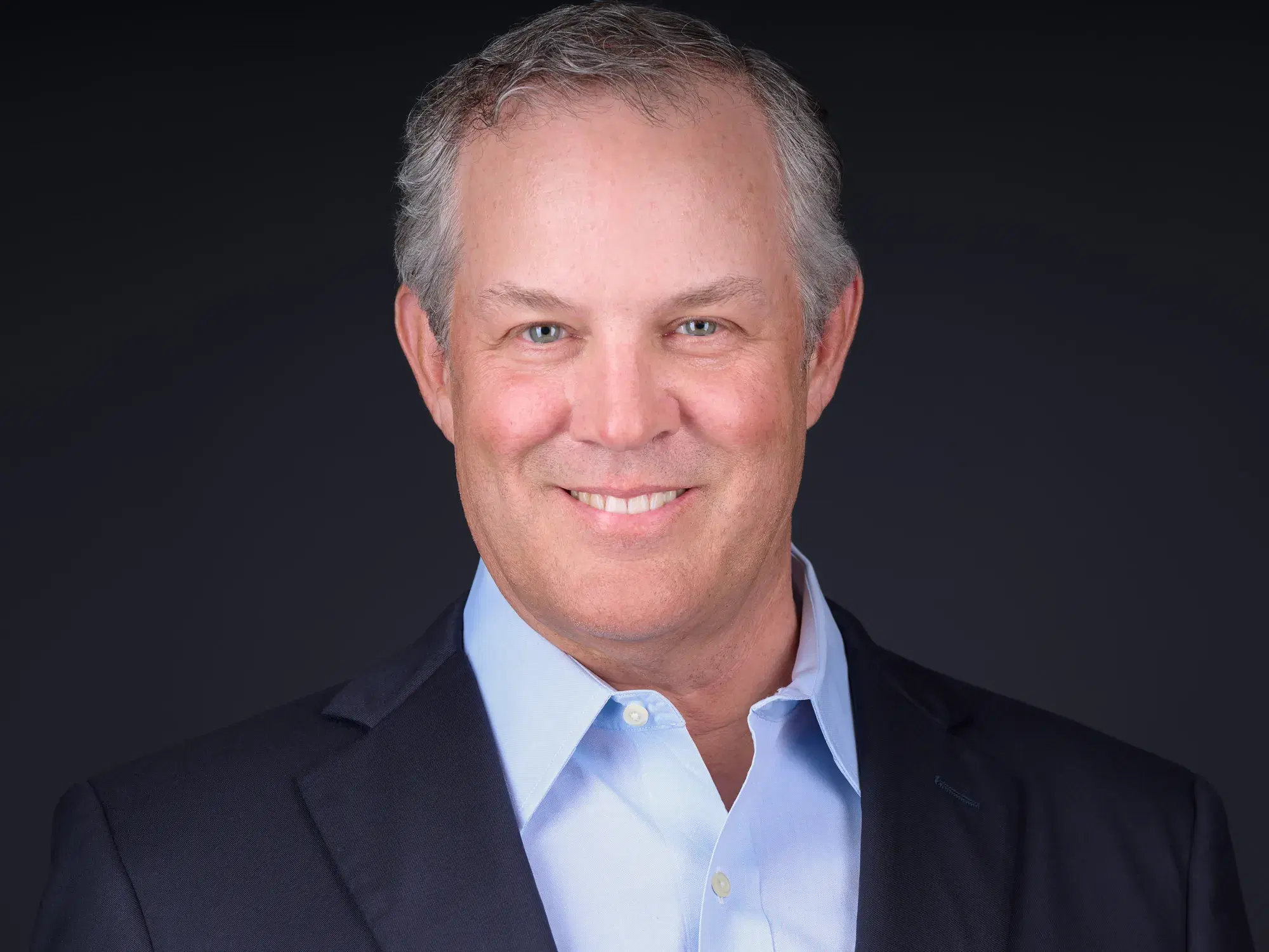 Man with silver hair in navy blazer and light blue shirt, warm smile against dark studio background — financial advisor headshot by Damon Bates Photography, Sherborn MA