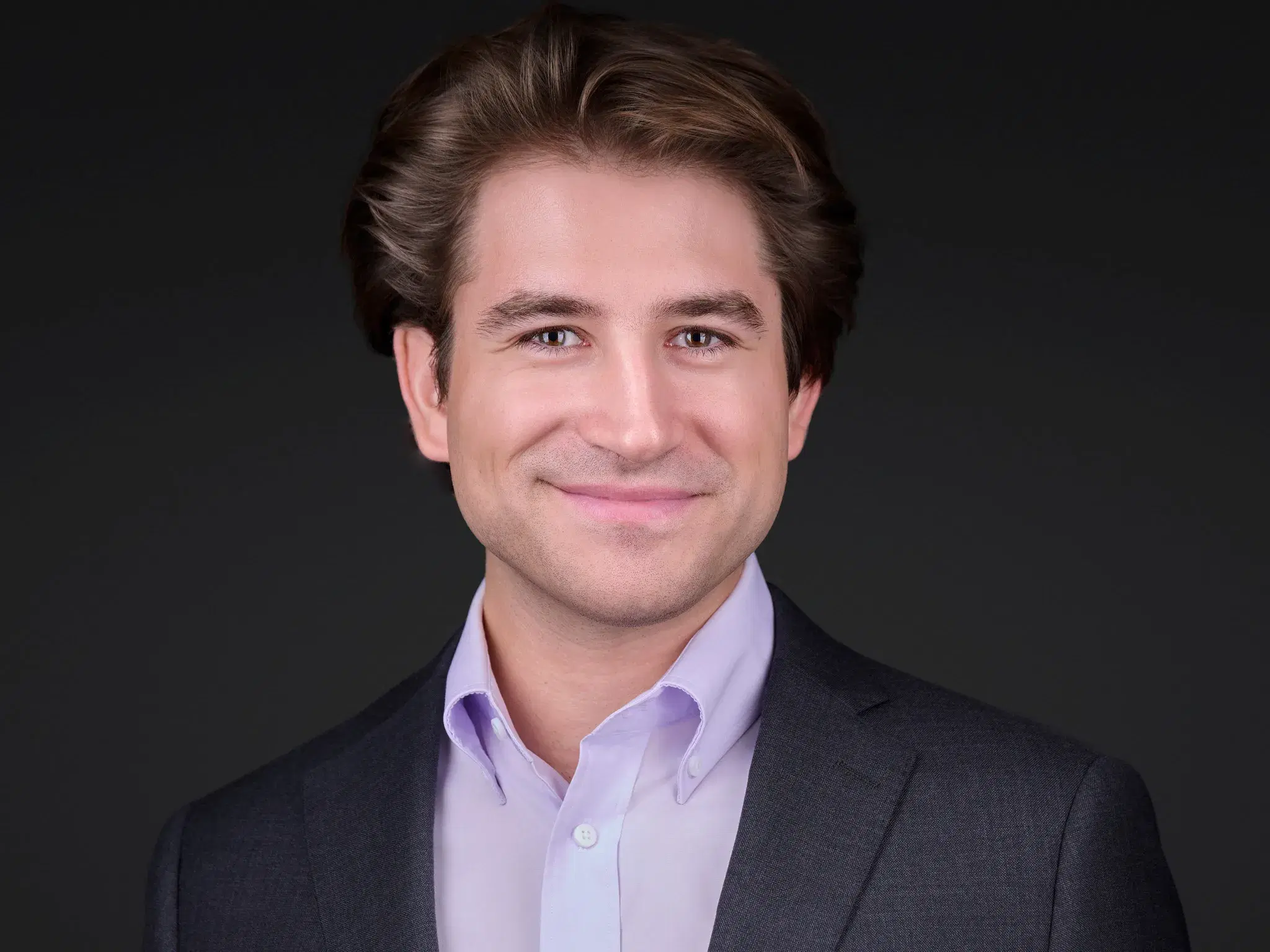 Professional headshot of a smiling man with wavy brown hair in a charcoal blazer and lavender shirt against a dark gray studio backdrop, by Boston team headshot photographer Damon Bates Photography