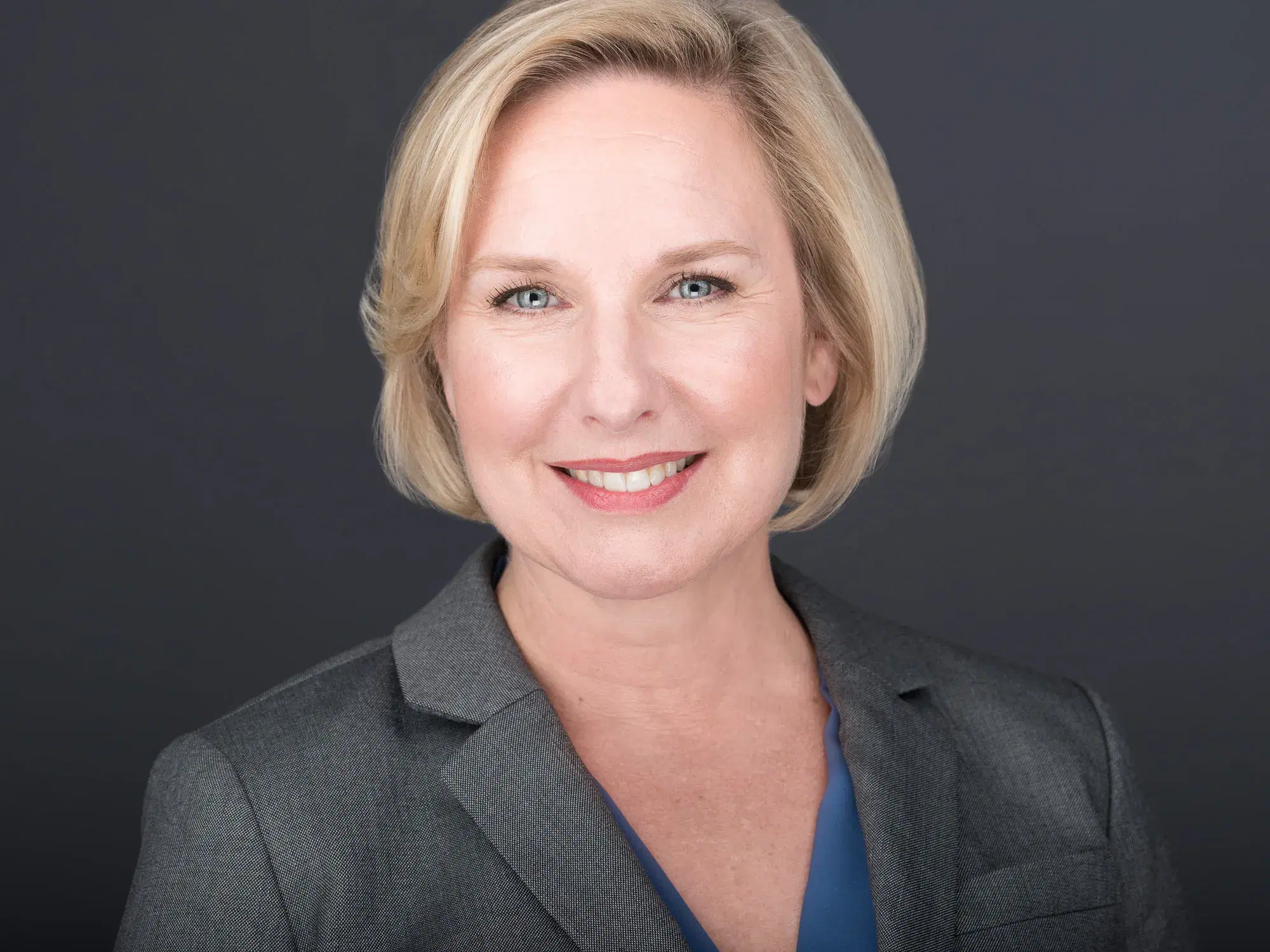 Corporate headshot of a woman with a blonde bob in a gray blazer, showing the consistent look of a team headshot day
